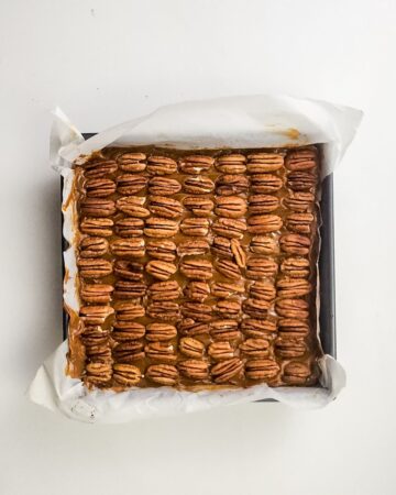 An 8x8 inch baking square pan with the layers to make millionaire brownies with the pecan layer showing.