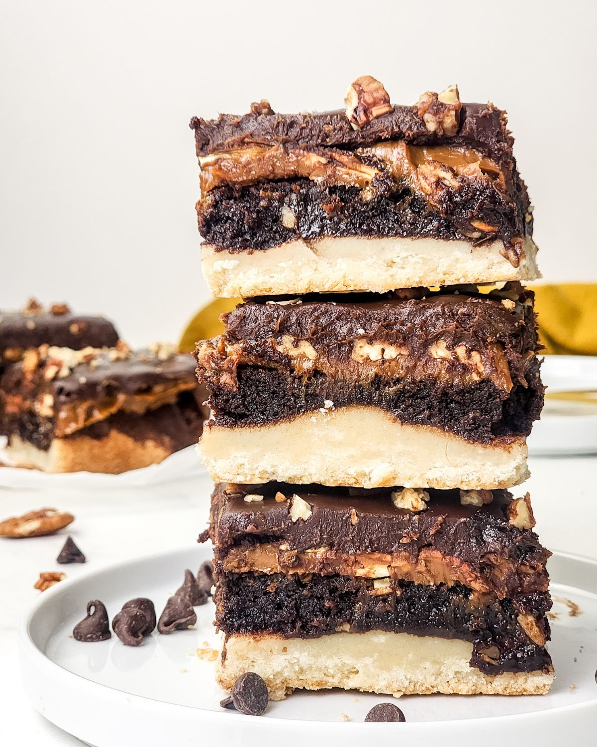 A stack of millionaire brownies on a white plate with chocolate morsels on the plate.
