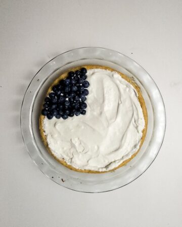 A glass pie plate with a sugar cookie base, whipped cream topping and blueberries in the left corner to make a fruit American flag pizza cookie.