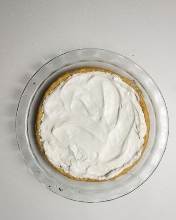 A glass pie plate with a sugar cookie base topped with whip cream topping to be the base of a fruit American flag pizza.