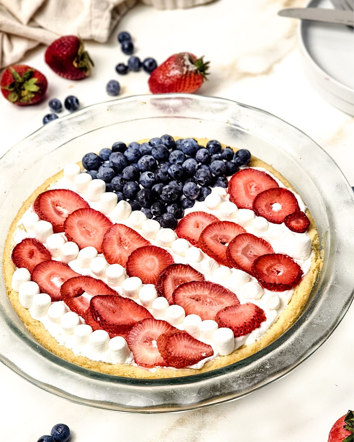 A glass plate with an American flag fruit pizza with a sugar cookie base topped with whipped cream and fruit to depict the American flag.