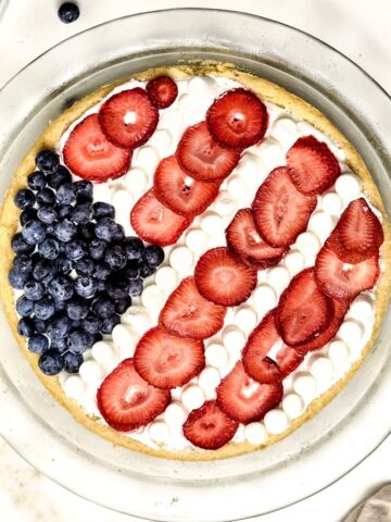 A glass clear pie plate with a sugar cookie base topped with whipped topping, blueberries, strawberries, and mini marshmallows to shape into the American flag.