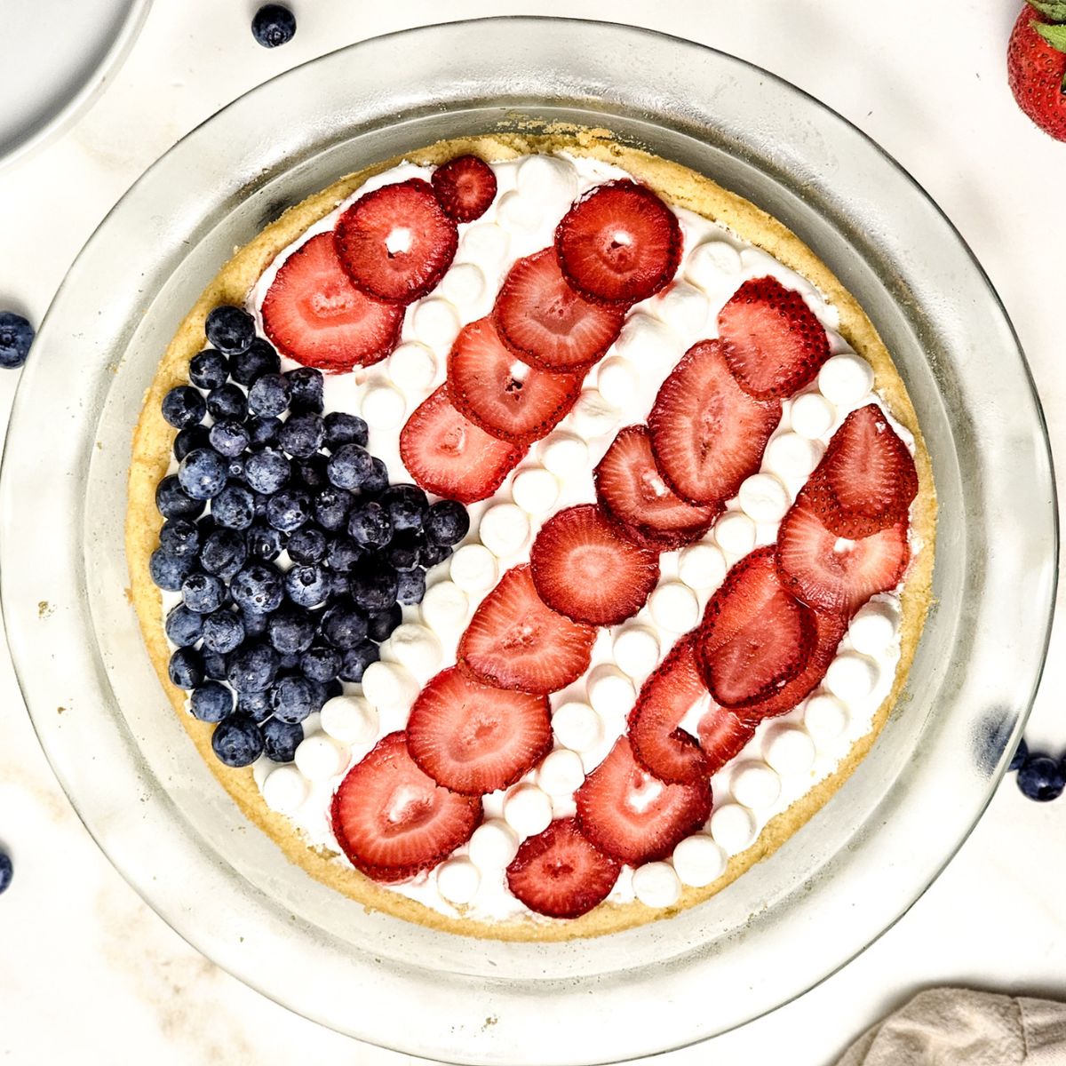 A glass clear pie plate with a sugar cookie base topped with whipped topping, blueberries, strawberries, and mini marshmallows to shape into the American flag.