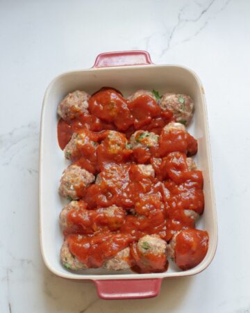 A large casserole dish with semi baked enchilada meatballs topped with enchilada sauce to make baked enchilada meatballs.