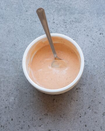 A white bowl with bang bang sauce in the bowl and a spoon in the bowl to drizzle on chicken skewers.