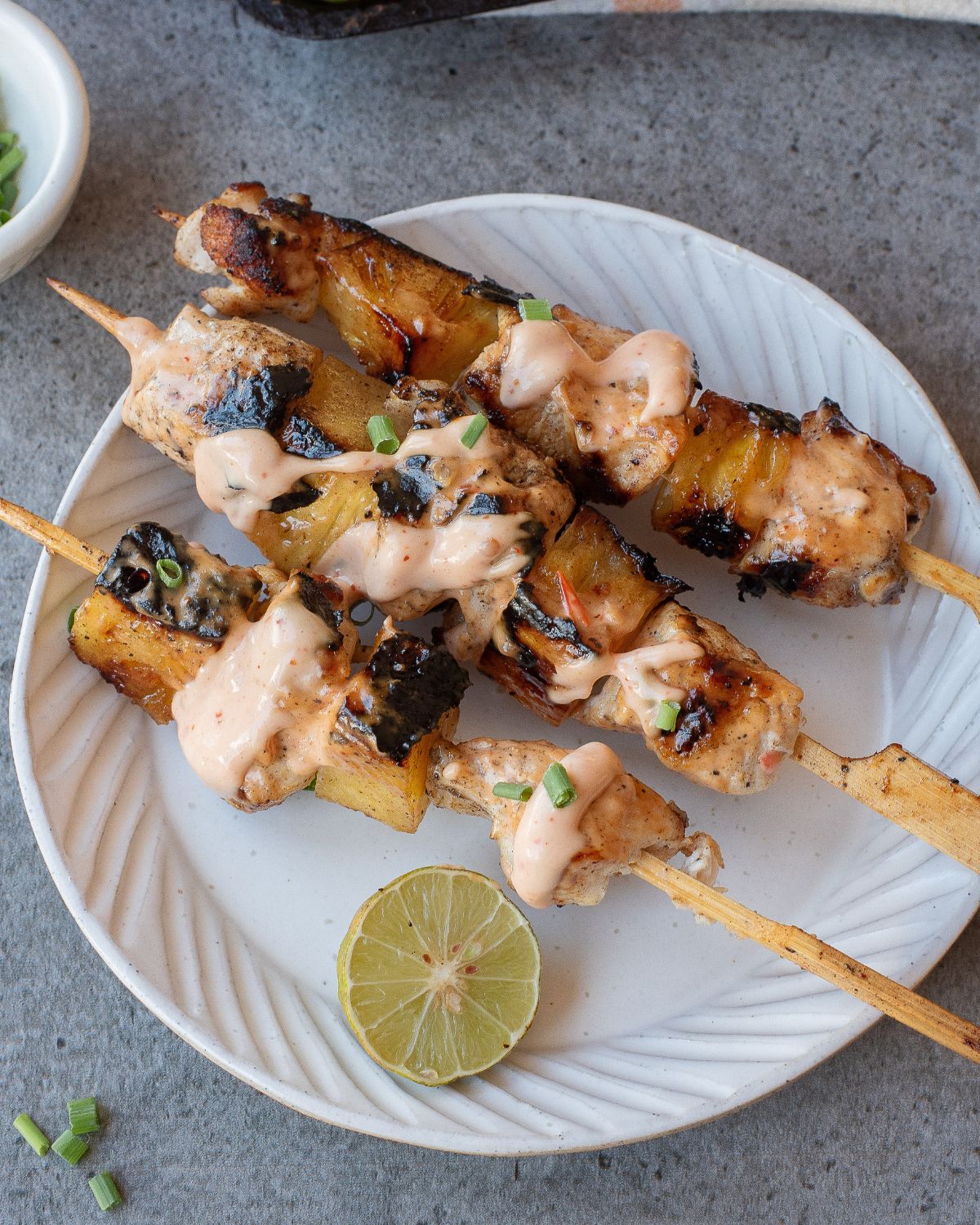 White plate with 3 chicken skewers with chicken kabobs and pineapple on the wooden skewers with bang bang sauce on the kabobs and a lime wedge.