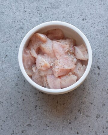 A white bowl with chicken cubes seasoned with salt and pepper to make bang bang chicken skewers.