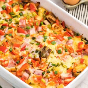 A white casserole dish with an egg casserole made with diced ham, peppers and cubed bread topped with sliced green onions.