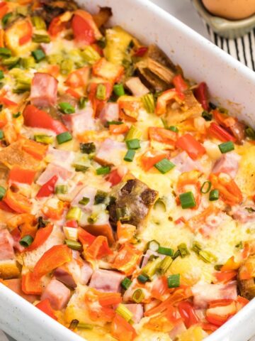 A white casserole dish with an egg casserole made with diced ham, peppers and cubed bread topped with sliced green onions.