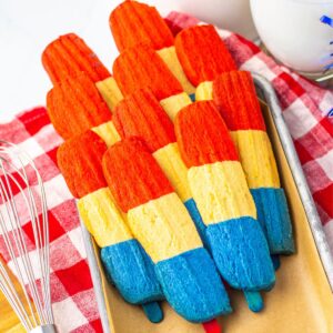 A tray of red, white and blue cookies shaped like rockets on a red and white checkered napkin with a whisk on the side of the tray of cookies.