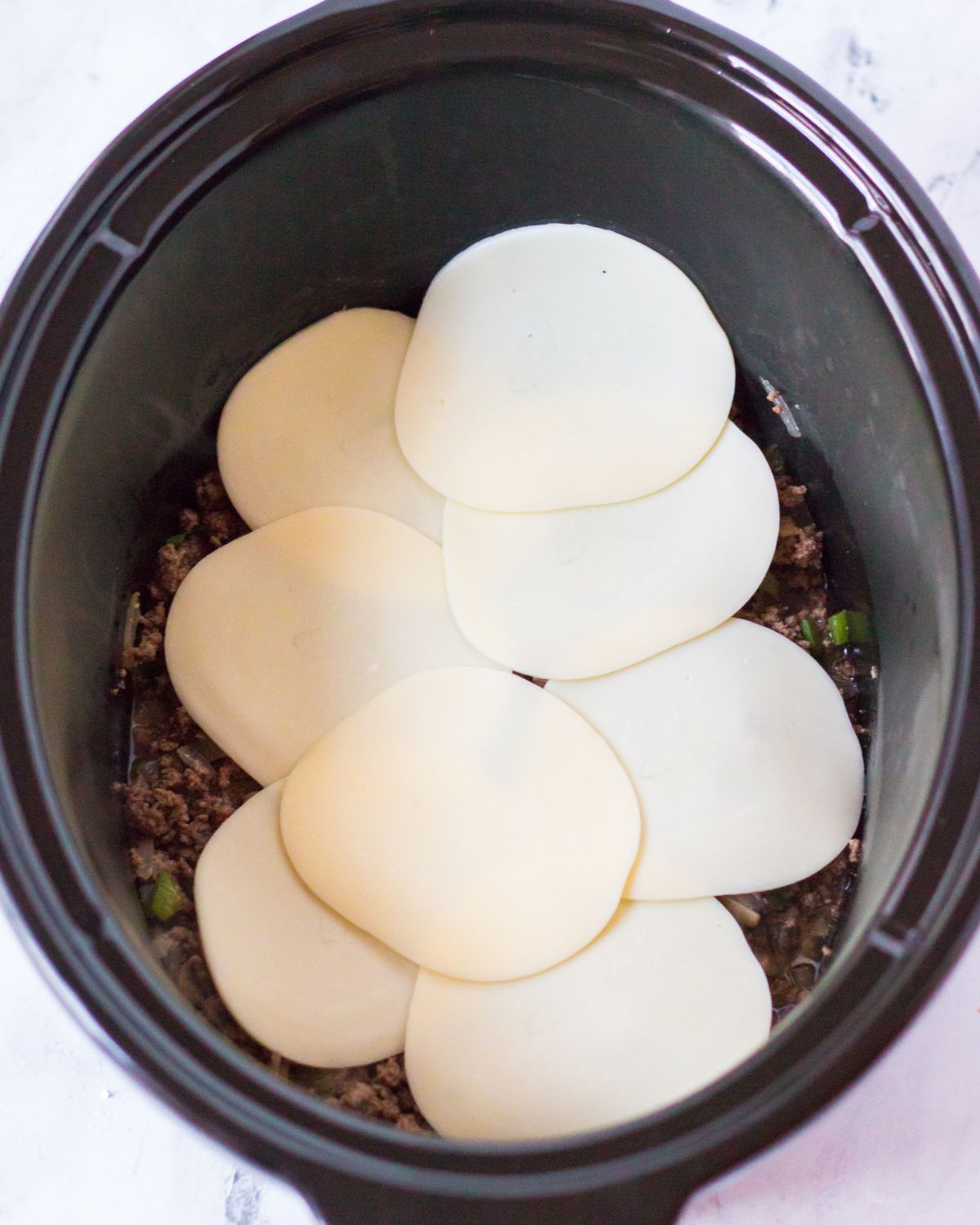 A crockpot of ground beef, peppers, and onions topped with slices of provolone cheese to make philly cheesesteak sloppy joes with ground beef.