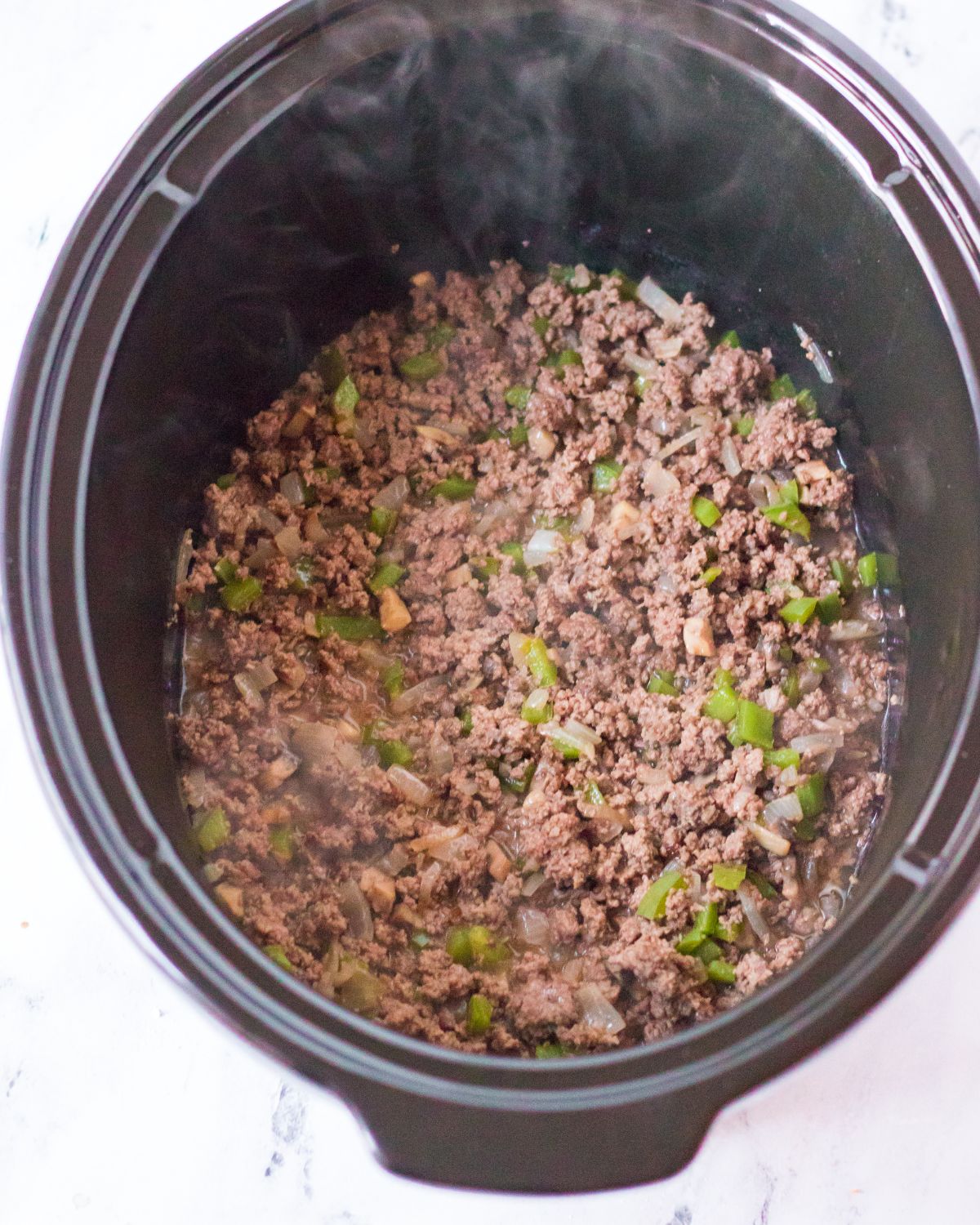A large brown slow cooker vessel with browned grouund beef, green peppers, and diced onions to make slow cooker philly cheesesteak sloppy joes.