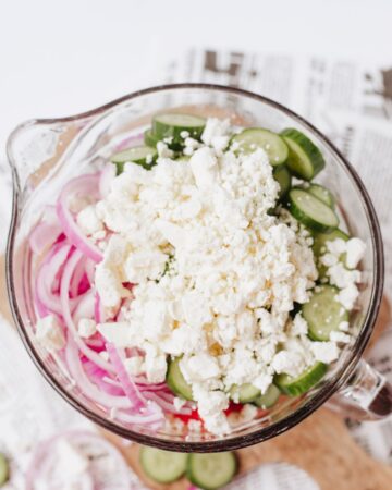 A glass mixing bowl with the prep ingredients of red onion, feta cheese, watermelon, and cucumbers in a bowl to make a salad.