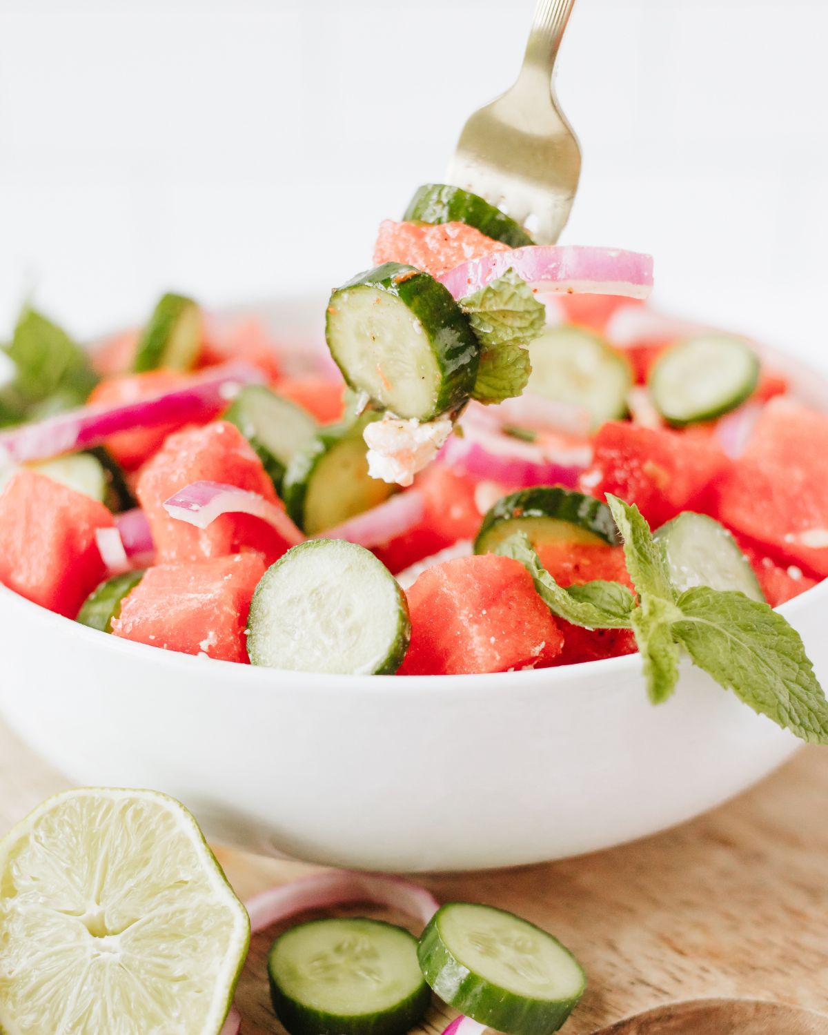 A white bowl with cubed watermelon, cucumber sliccesm and mint leaves in a bowl.