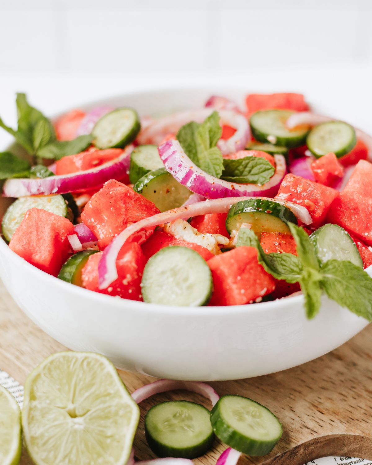 A white salad bowl filled with sliced cucumbers, sliced red onions and cubed watermelon in a citrus vinaigrette.