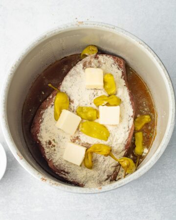 A pot roast in the instant pot vessel with butter and banana peppers on the roast