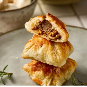 A stack of cooked puff pastry. The top puff pastry is sliced open showing beef tenderloin on a grainy mustard and mushrrom filling.