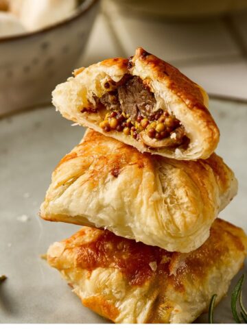 A stack of cooked puff pastry. The top puff pastry is sliced open showing beef tenderloin on a grainy mustard and mushrrom filling.