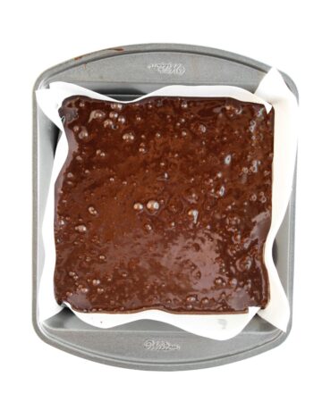 A square baking pan lined with parchment paper filled with brownie batter to make pumpkin browies with a cream cheese swirl.