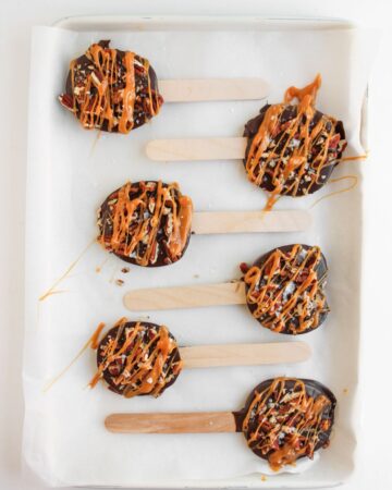 A tray with wax paper on the baking tray with apple slices dipped in chocolate with pecans and drizzled caramel on wooden sticks.