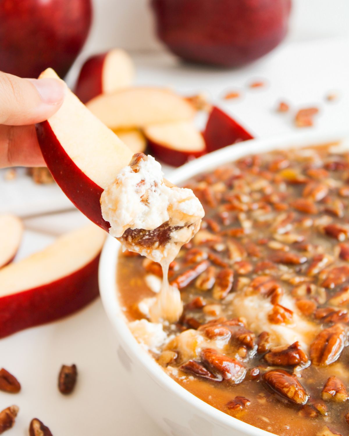A white bowl with a cream cheese base dip topped with a pecan pie topping and an apple being dipped into the dip.