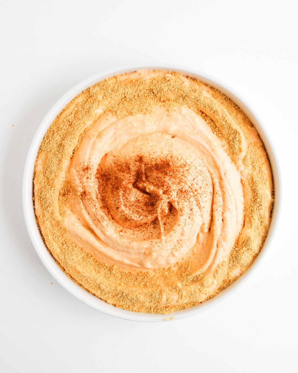 A white bowl with a no bake pumpkin pie dip topped with graham cracker crumbs and ground cinnamon.