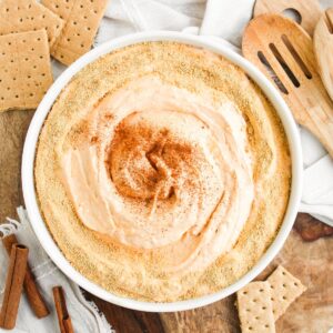 A bowl of no bake pumpkin pie dip with graham cracker crumbs and a sprinkle of cinnamon on top. GRaham crackers are scattered on the side of the bowl with cinnamon sticks.