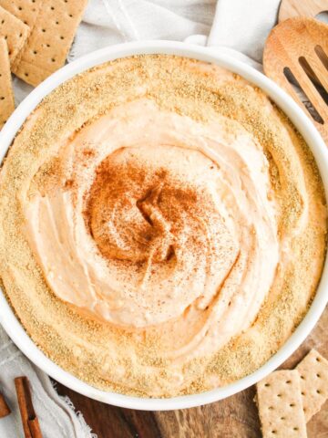 A bowl of no bake pumpkin pie dip with graham cracker crumbs and a sprinkle of cinnamon on top. GRaham crackers are scattered on the side of the bowl with cinnamon sticks.
