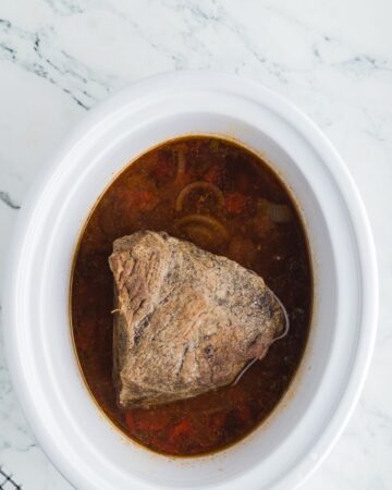 Slow cooker vessel with a browned rump roast, onions, fire roasted tomatoes and seasonings to make Mexican shredded beef.