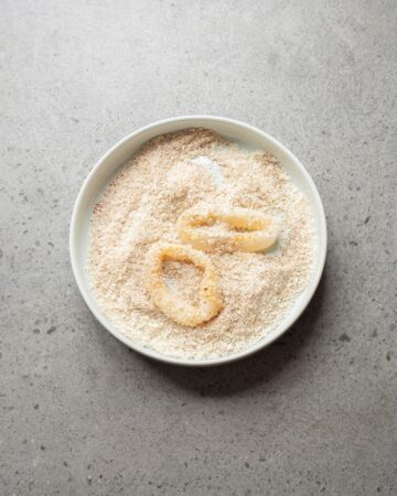 A white bowl with panko breadcrumbs and rings of calamari in the bowl to be breaded.