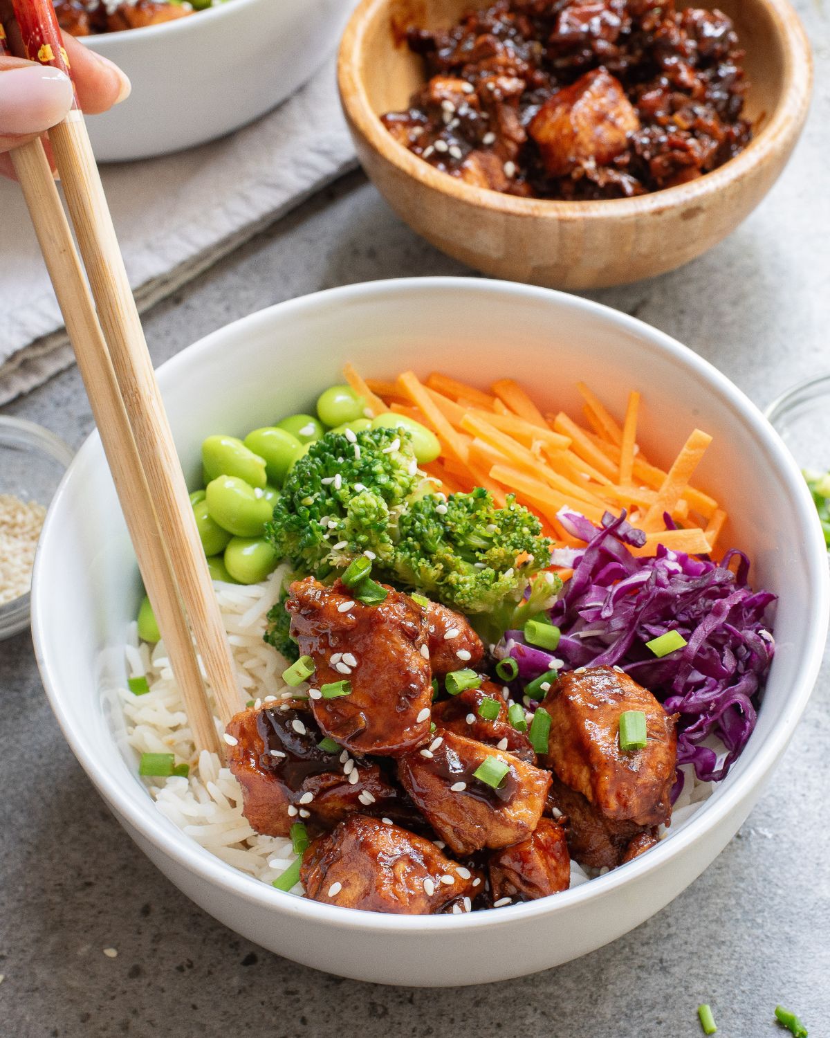A white bowl with white rice, teriyaki chicken, shredded carrots, broccoli, edamame and red cabbage