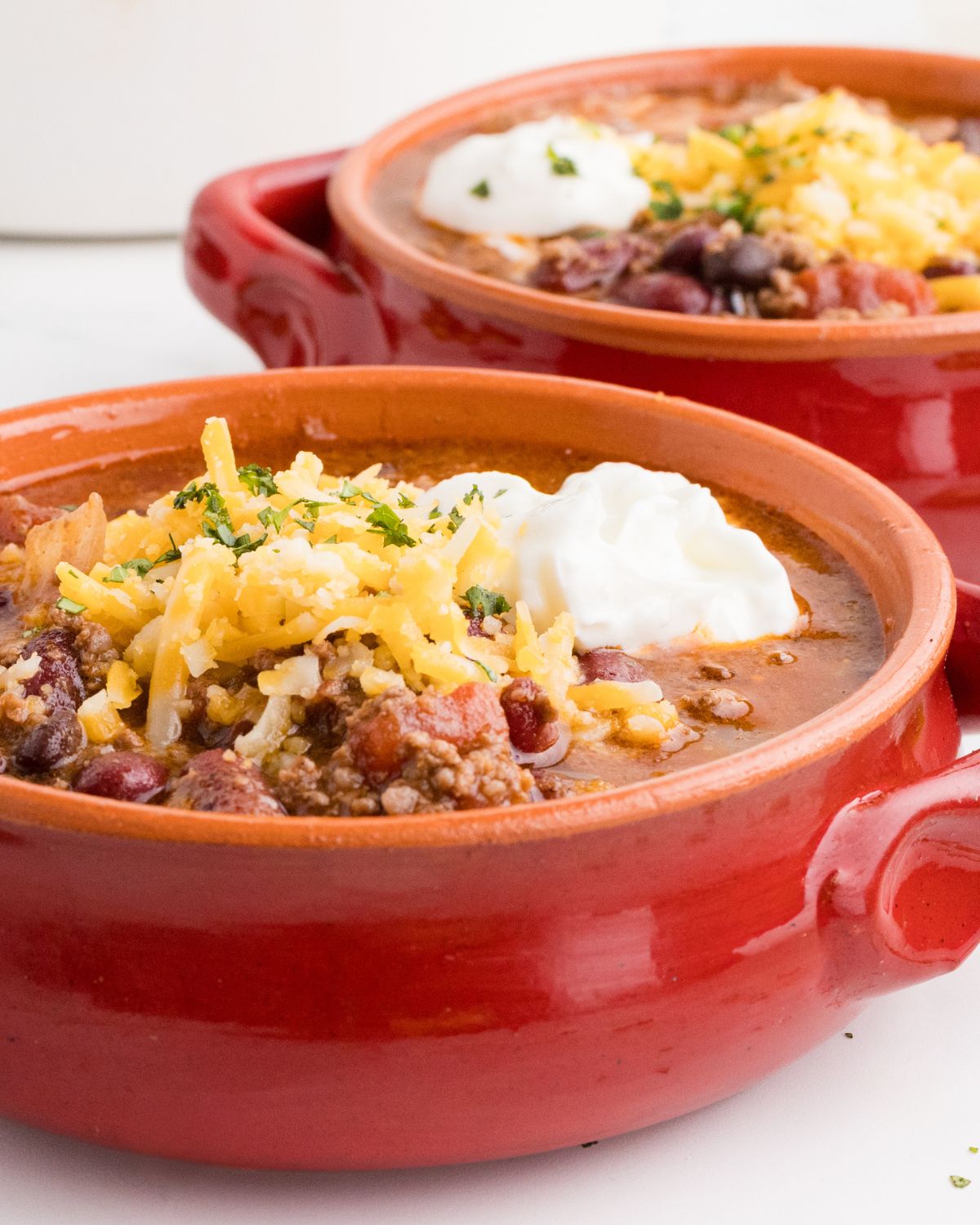 Two ceramic red bowls with beef chipotle chili in the bowls topped with shredded cheddar cheese, sour cream, and chopped parsley.