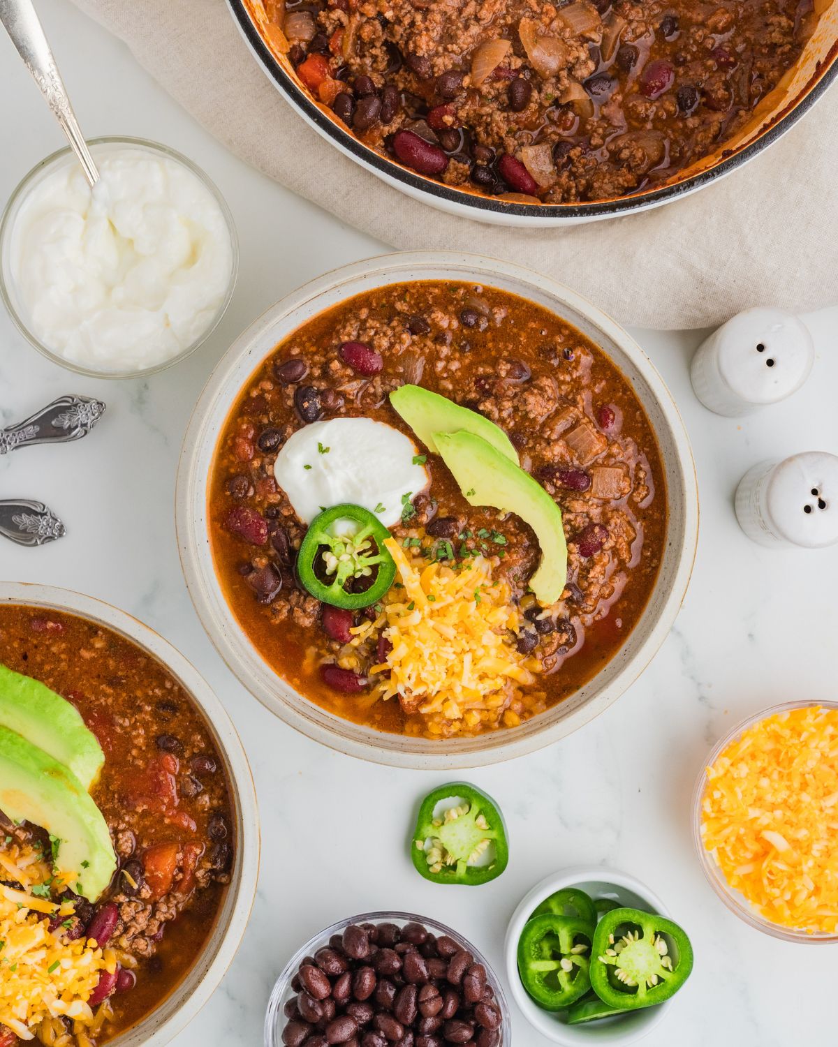 Smoky chipotle chili with chipotle peppers in a bowl topped with jalapeno slice, shredded cheddar cheese, avocado, and sour cream with bowls of ingredients surrounding the bowl.