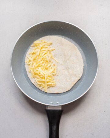 A saute pan with a tortilla topped with shredded cheddar cheese.