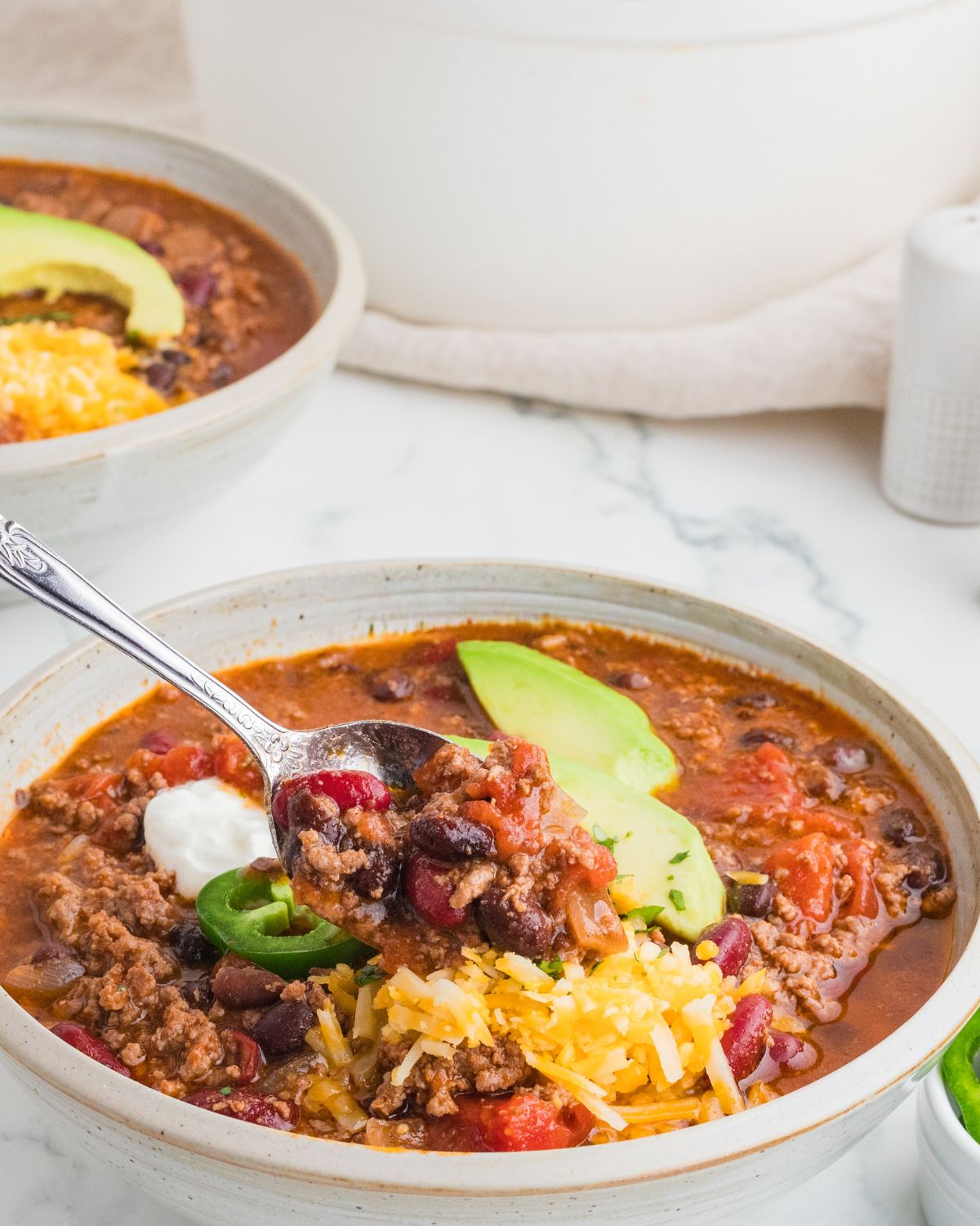 A white ceramic bowl with ground beef chipotle chili topped with shredded cheese, avocado, and sour cream. With a spoon picking up a scoop of the chipotle chili.