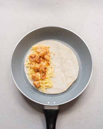 A saute pan with an tortilla in it, topped with shredded cheddar cheese and seasoned sauteed shrimp.