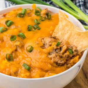 A white serving bowl with chili cheese dip and a taco chip in the dip.