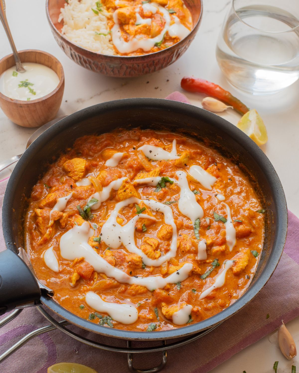 Indian inspired butter chicken recipe in a saute pan with a bowl of basmati rice and butter chicken behind the pan.