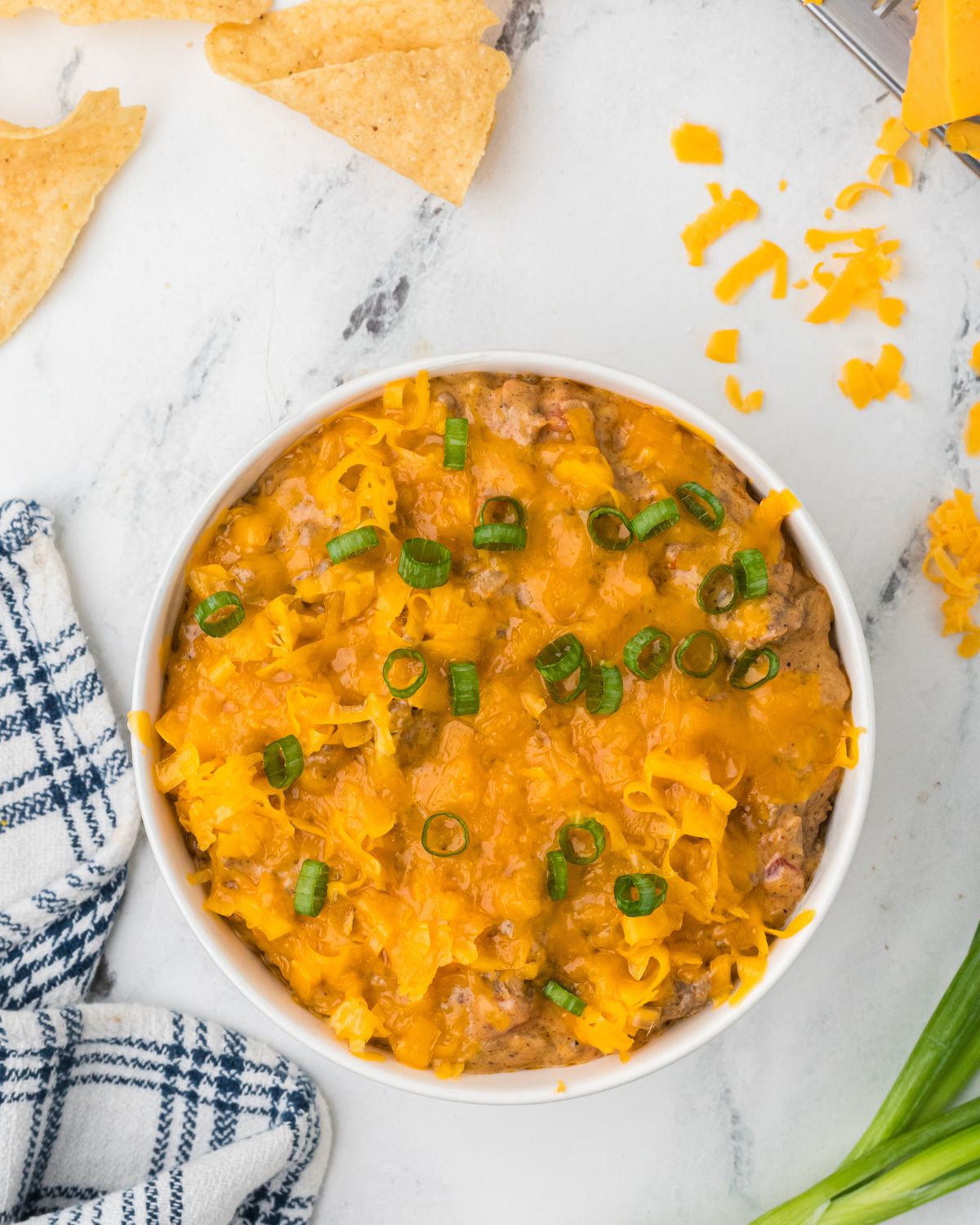 A white bowl with chili cheese dip topped with shredded cheese and diced green onion.