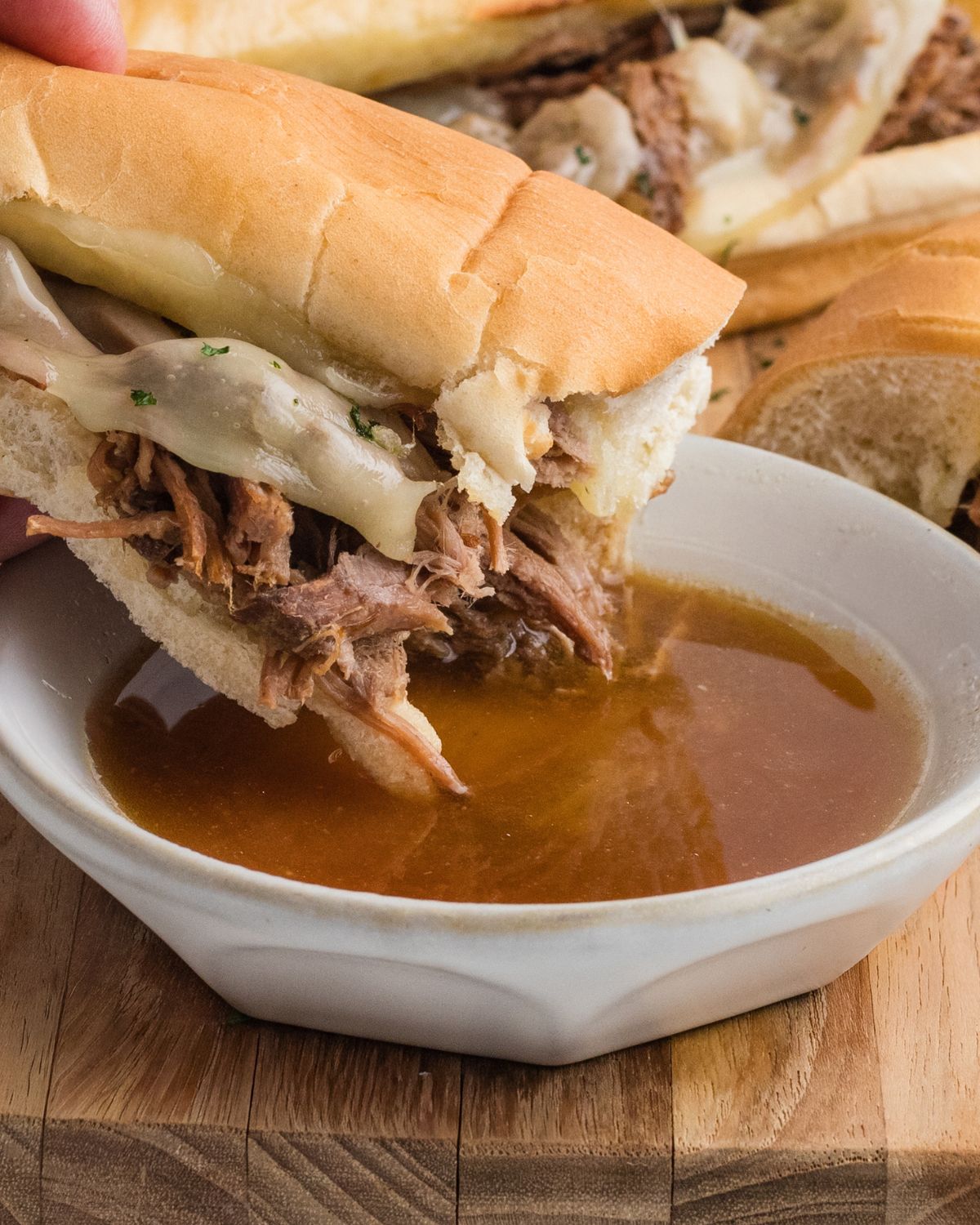 Crockpot shredded french dip beef on hoagie bread with provolone cheese dipped into au jus beef in a white bowl.