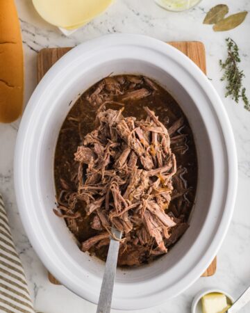 A white slow cooker vessel with a rump roast, beef broth, soy, and onions slow cooked to make au jus french dip sandwiches in the crockpot.