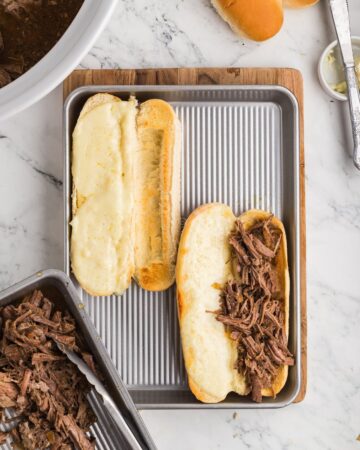 A silver tray wth a hoagie rolls with provolone cheese and shredded beef for slow cooker beef french dip sandwiches