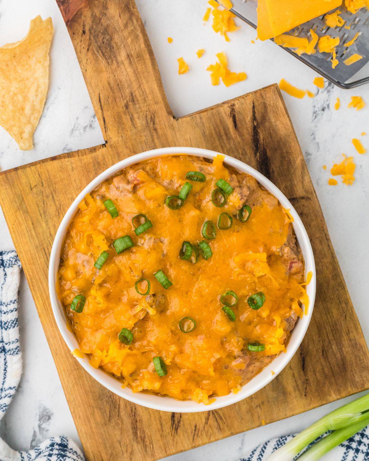 A wooden cutting board with a white bowl with chili cheese dip topped with green onions. 
