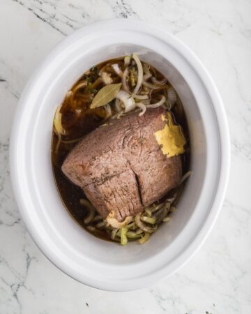 A slow cooker vessel with browned roast, sliced onions, and broth to make French Dip Sandwiches in the crockpot.