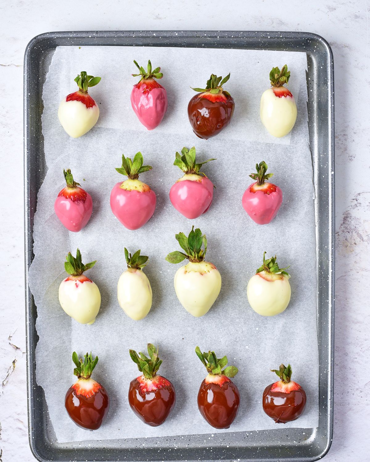 A baking sheet layered with parchment paper with fresh strawberries dipped in milk chocolate colored in white, red, and chocolate to make chocolate covered strawberries.