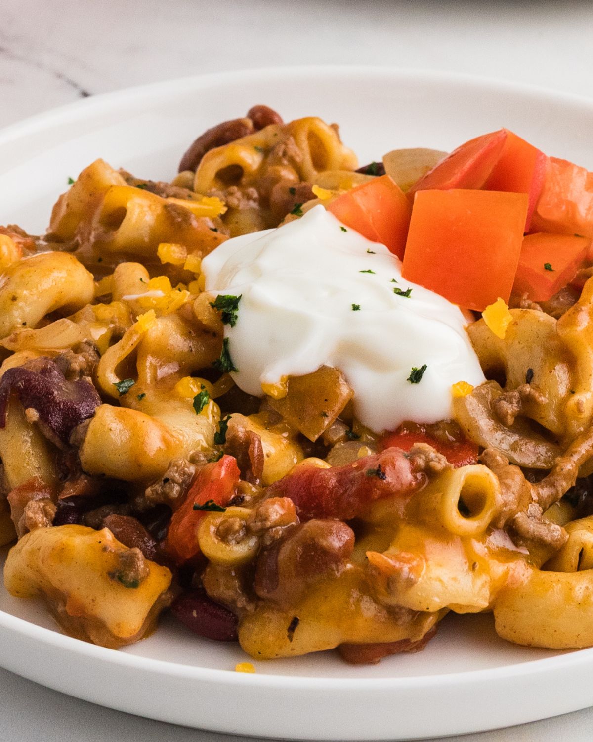A recipe for chili mac and cheese topped with sour cream and chopped tomatoes on a white plate.