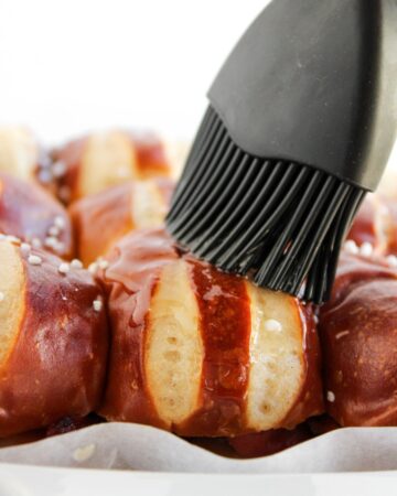 A silicone brush brushing maple syrup on pretzel buns for breakfast sliders.