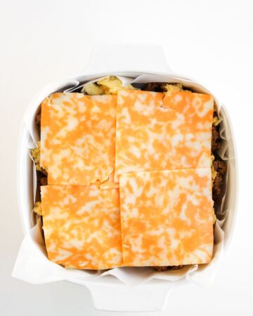 The construction of breakfast sliders on the bottom half of slider rolls with eggs, bacon, sausage, and cheese slices.