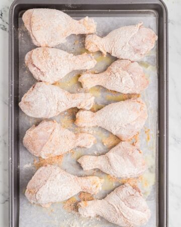 Chicken drumsticks in seasoned flour on a baking sheet to make baked crispy chicken.