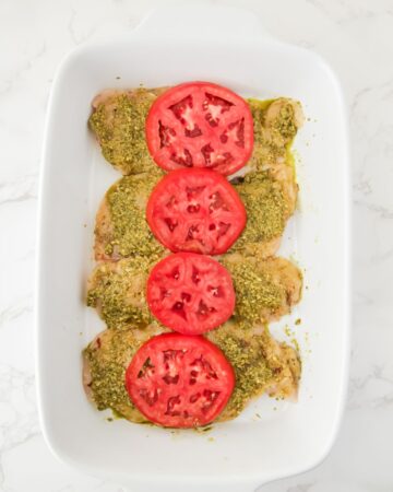 A baking dish with chicken topped with pesto and a slice of tomatoes to make the step by step pesto chicken bake.